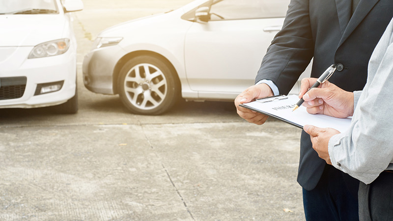 Two formally dressed professionals inspecting and appraising two vehicles—one holding a clipboard and pen while the other records details