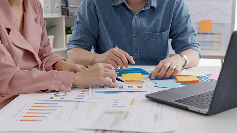 Two professionals working together analyzing business data, charts, and graphs on a table with a laptop and documents